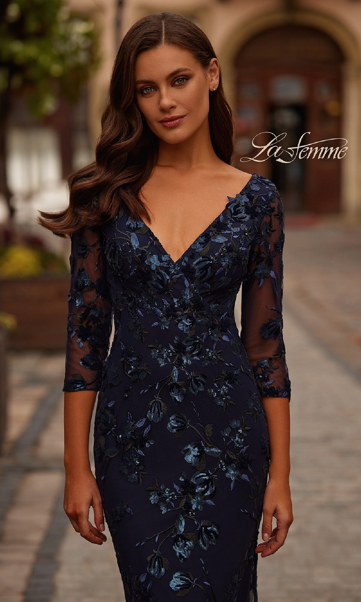 A woman wearing a Navy Blue Sequin-Lace Formal Dress—La Femme 31836—stands on a cobblestone street, with the dress’s floral embroidery and sheer sleeves elegantly displayed.